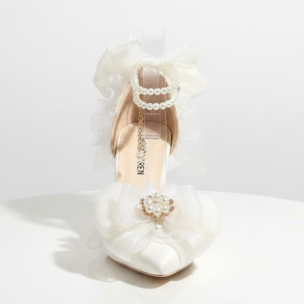 A single embellished high-heeled shoe displayed on a gray surface with decorative props. The shoe is predominantly white with sheer white ruffled bows, pearl-like bead trim, and a small gold-tone chain detail.