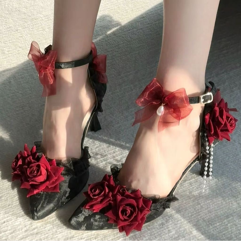 Black lace high-heeled shoes decorated with red roses and red sheer ribbon bows, shown on a model's feet against a gray concrete background.