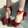 Black lace high-heeled shoes decorated with red roses and red sheer ribbon bows, shown on a model's feet against a gray concrete background.