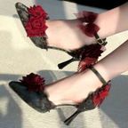 A pair of pointed-toe high heels worn on feet, featuring dark lace uppers with red fabric roses and red sheer bow accents on the ankle straps, photographed outdoors on a light textured surface.