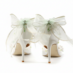 A pair of white high-heeled shoes decorated with large sheer white bows, delicate gold trim, and small green floral accents, displayed on a soft white surface with gold confetti.