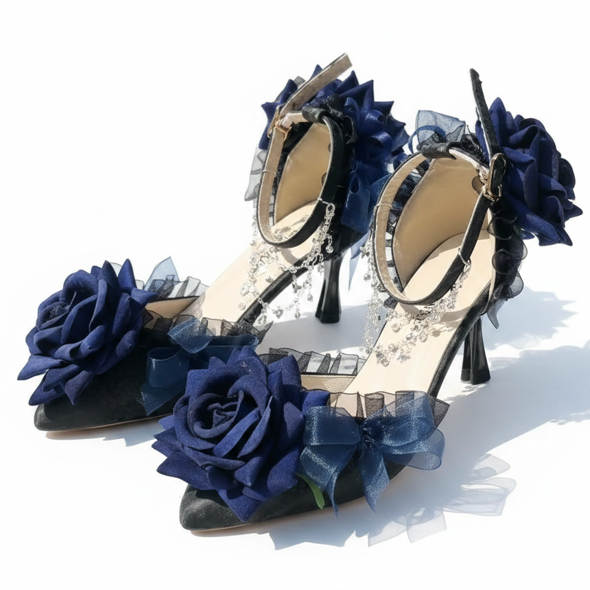 A pair of black high-heeled sandals decorated with large navy blue fabric roses, sheer navy bows, and silver rhinestone chain details, displayed on a white textured blanket outdoors.