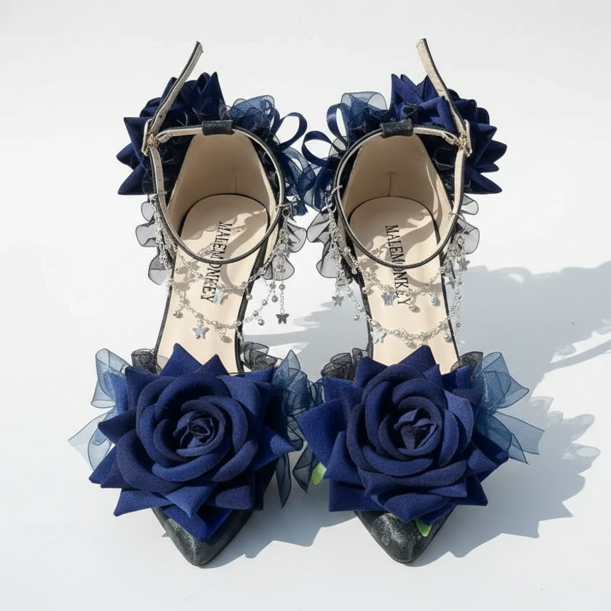 A pair of pointed-toe heeled sandals with dark navy blue fabric roses, sheer tulle, and silver rhinestone trim, displayed on a textured white surface.