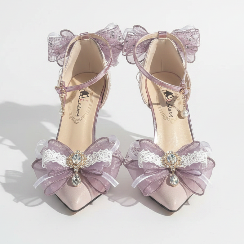 A pair of pointed-toe high heels in a pale blush pink with lavender-purple ribbon bows, white lace trim, and rhinestone embellishments, displayed on a white draped fabric with a decorative silver tray.