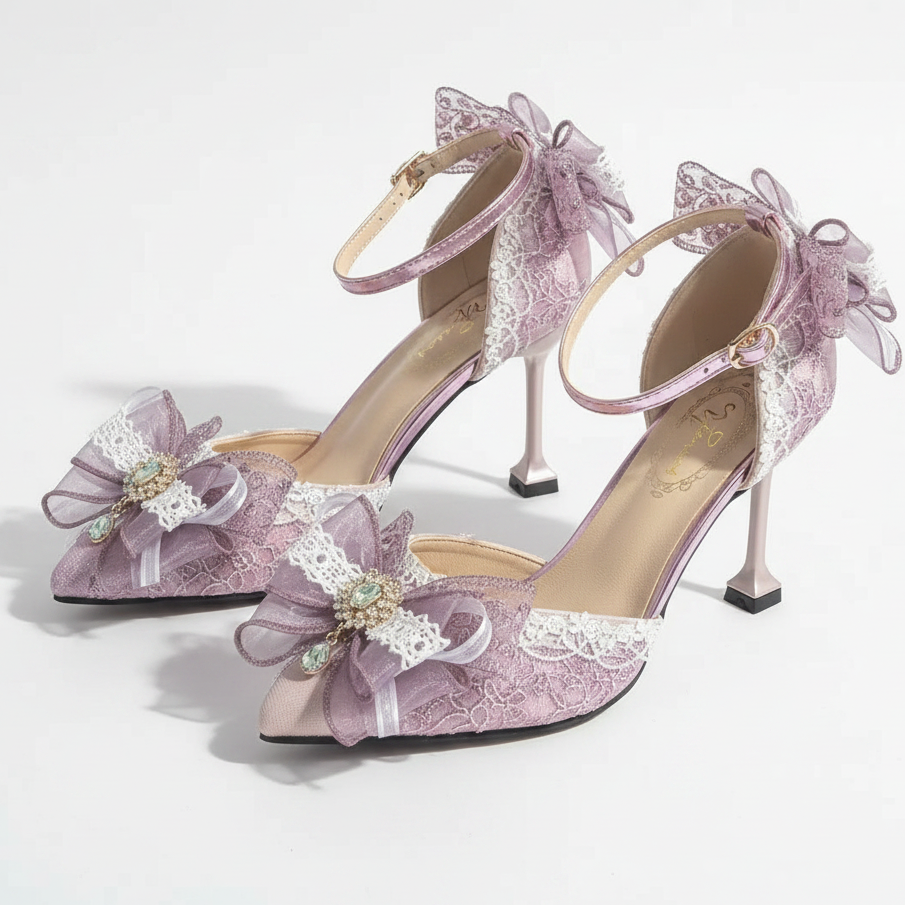 A pair of women's high-heeled sandals in a pale lavender satin, decorated with white lace, sheer purple ribbon bows, and small pearl-like accents. The shoes are displayed on a white surface with soft shadows and a decorative white object in the background.
