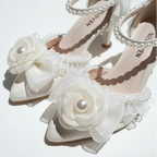 A pair of white satin high-heeled sandals decorated with large fabric roses, pearl-like center beads, and a border of small pearls, displayed on a white ruffled fabric background.