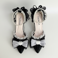 A pair of black and white bridal-style pointed-toe flats with ruffled trim, pearl bead straps, and large bow details, arranged on a white fabric surface with pink flowers and lace props. The insole shows the brand name FALINUKA.