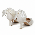 A pair of off-white satin high heels with large bows, pearl bead details, and low block heels, displayed on a textured beige surface against a warm beige background.