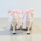 A pair of white lace high-heeled shoes decorated with large light pink ribbon bows, dangling pearl-like beads, and black-and-white striped heel tips, photographed on a light surface with a blurred indoor background.