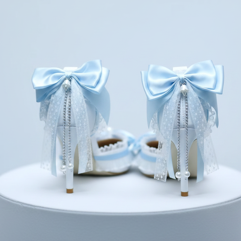 A pair of white high-heeled shoes decorated with large light blue satin bows, sheer white lace ribbons, pearl-like beads, and dangling silver chains, displayed on a round white pedestal against a pale blue background.