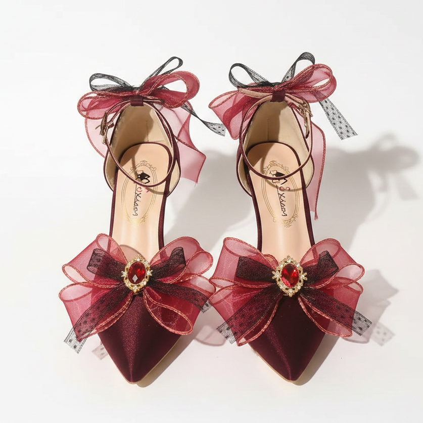 A pair of high-heeled pointed-toe sandals with large burgundy and dark red sheer bow embellishments on the toes and ankles, accented with rhinestone details, shown on a reflective surface.