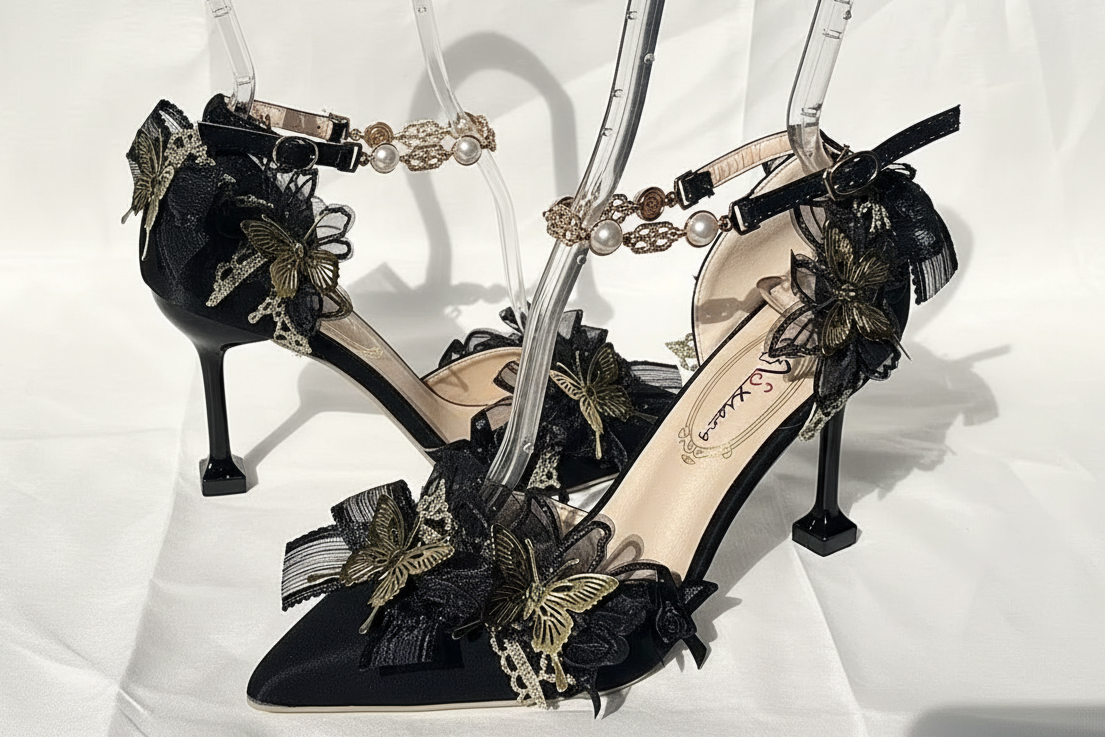 A pair of black pointed-toe high heels with ankle straps, decorated with black lace ruffles, gold butterfly embellishments, and pearl-like bead details, displayed on clear stands against a white fabric backdrop.