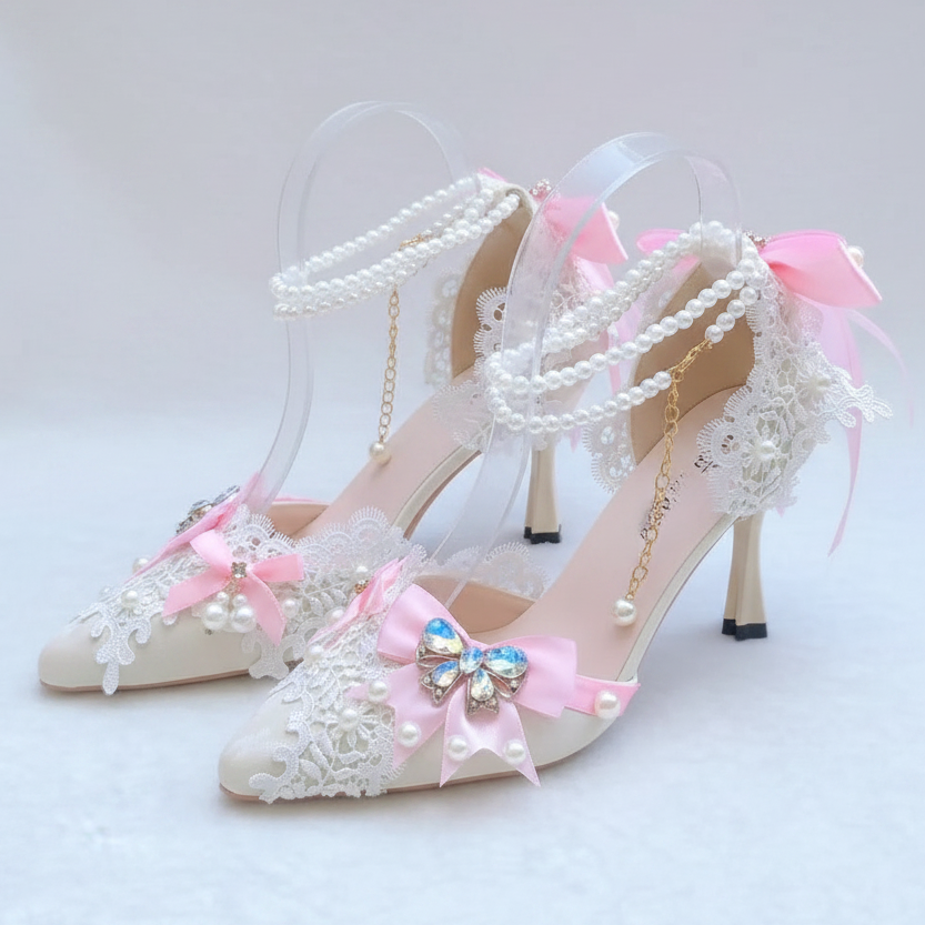 A pair of bridal-style high heels with clear straps, white lace, pink satin bows, pearl bead details, and a jeweled bow on the toes, displayed on a white surface with a blurred garden background.