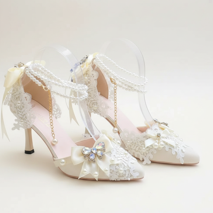 A pair of cream-colored pointed-toe high heels decorated with lace, pearl straps, clear acrylic arches, and a blue crystal bow, displayed on a white surface with a blurred garden background.