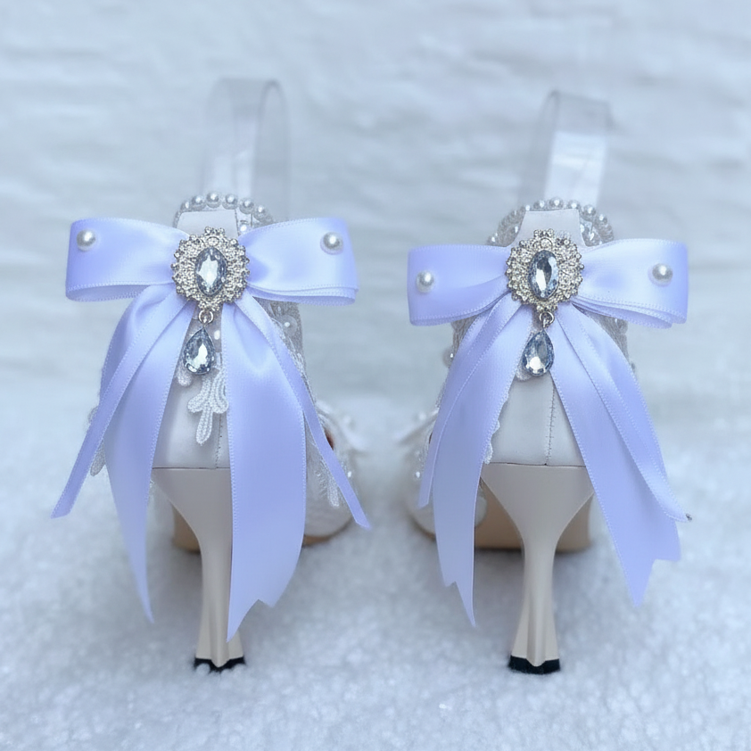 A pair of bridal high heels displayed on clear stands, decorated with large light lavender satin bows, pearl-like beads, and a central jeweled brooch. Pink flowers are blurred in the background against a white textured backdrop.