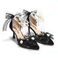 A pair of black lace high-heeled sandals with pearl ankle straps, sheer black tulle bows, and white floral embellishments on a gray marble-like surface.