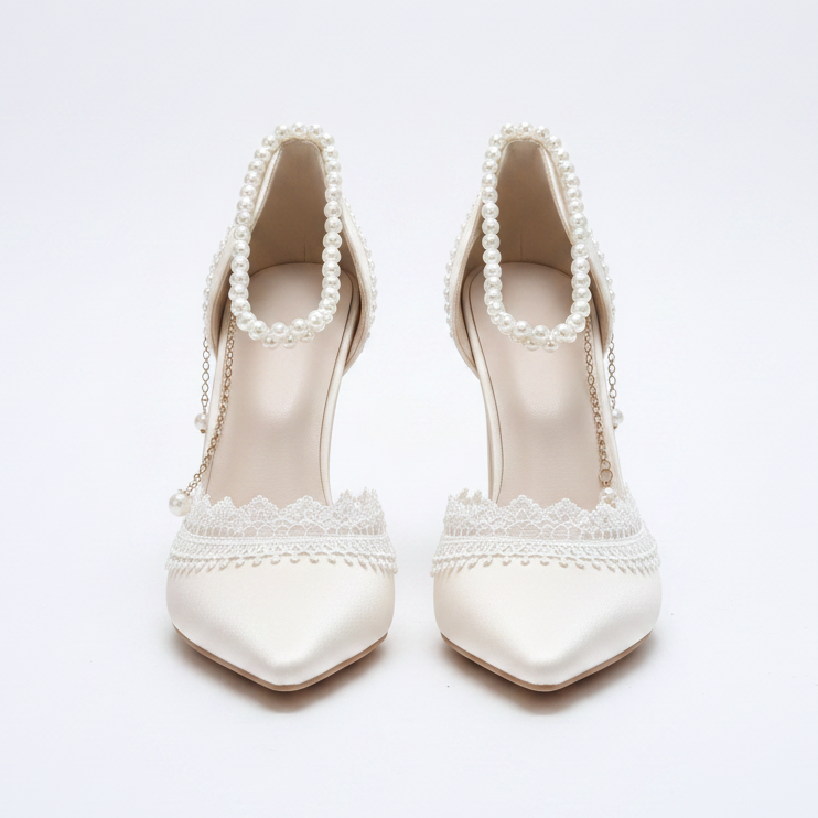 A pair of pointed-toe high heels in a warm ivory satin with pearl bead trim, lace detailing, and delicate chain straps, displayed on an ornate gold mirror tray.