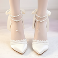 A pair of pointed-toe high heels worn on feet, featuring cream satin uppers, lace trim, pearl ankle straps, and large ribbon bows, shown against a soft light background.