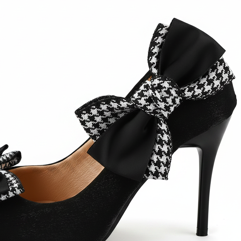 Black high-heeled pump shoes with a large black-and-white houndstooth bow on the back strap, shown on a beige background with one shoe in the foreground and another partially visible behind it.