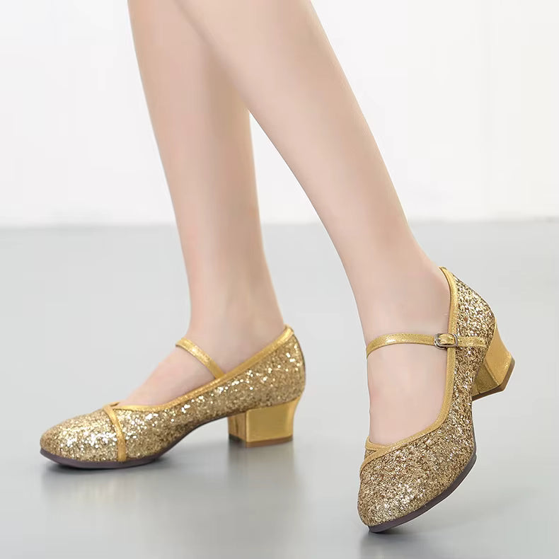 A pair of women's glittery gold dance shoes with low block heels and Mary Jane straps, shown on a model's feet against a neutral studio background.