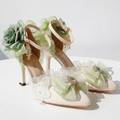 A pair of embellished high-heeled sandals with pale green satin roses, white lace butterflies, and faux succulent details, displayed on a light surface with decorative shadows.