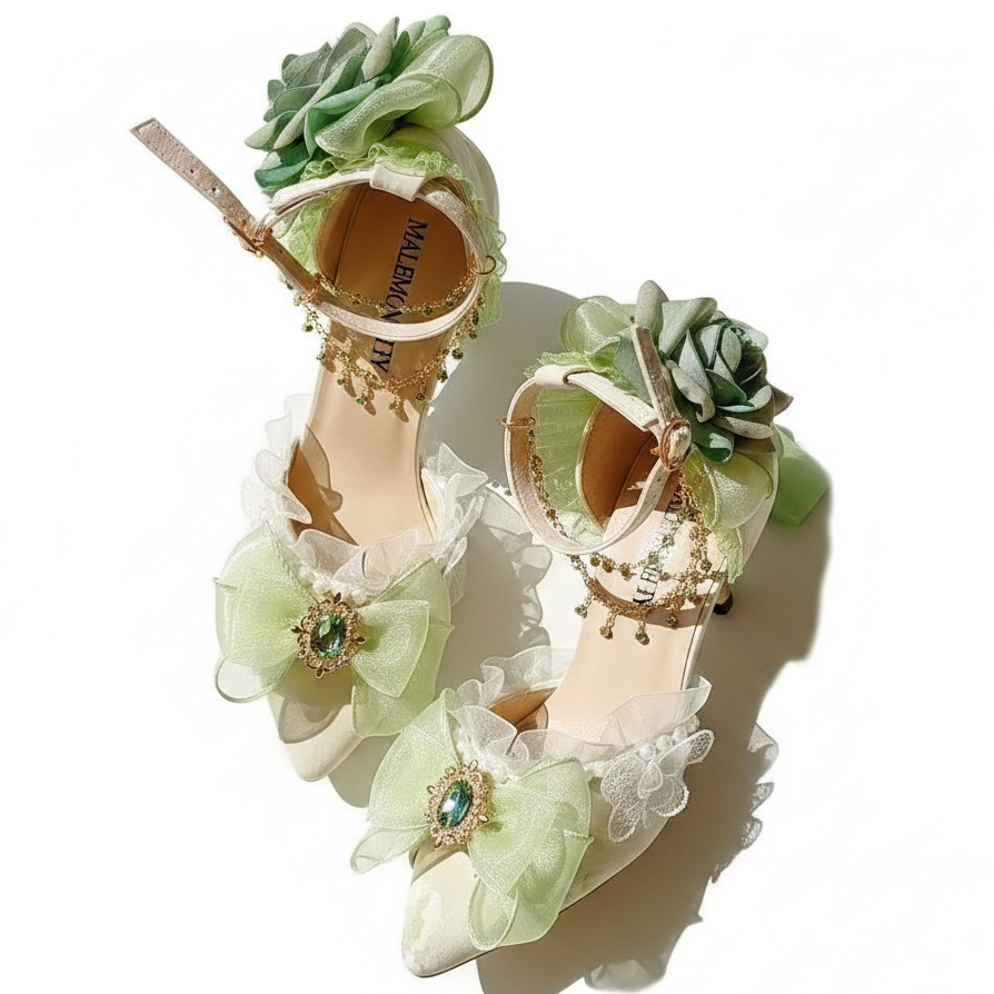 A pair of ornate beige high-heeled sandals decorated with pale green ruffled fabric flowers, lace trim, and jeweled embellishments, arranged on a light surface with a decorative mirror in the background.