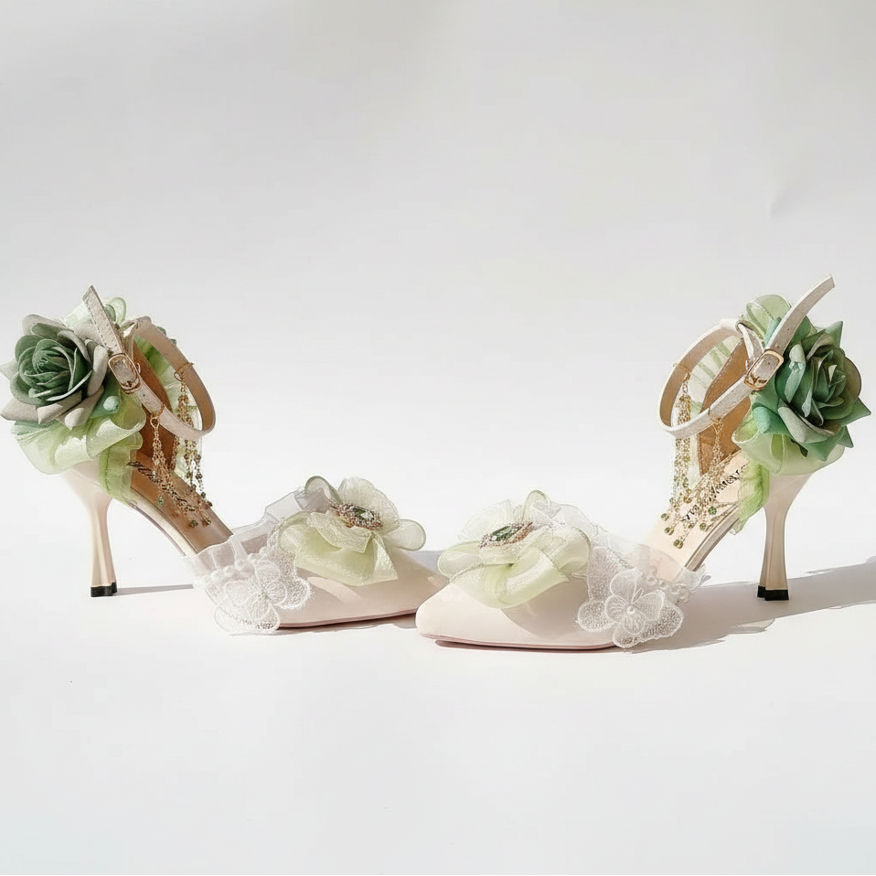 A pair of beige high-heeled sandals decorated with large green fabric flowers, white lace appliqués, and dangling gold-toned embellishments, displayed on a white surface with a soft floral backdrop.